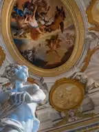 A historical building with two towers, surrounded by manicured gardens, statues, and ornate stone columns under a clear blue sky.