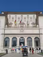 A historical building with two towers, surrounded by manicured gardens, statues, and ornate stone columns under a clear blue sky.