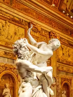 A historical building with two towers, surrounded by manicured gardens, statues, and ornate stone columns under a clear blue sky.