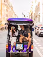 A historical building with two towers, surrounded by manicured gardens, statues, and ornate stone columns under a clear blue sky.