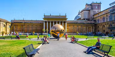 Tourists relax on benches in a courtyard with a large bronze sculpture and historic building under a clear blue sky.