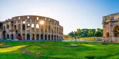 Ancient Colosseum and Arch of Constantine in Rome, with green grass and scattered visitors under a clear blue sky.