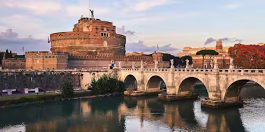 Historic stone bridge with arches spans a river, leading to a large cylindrical castle. Autumn trees and buildings in the background.