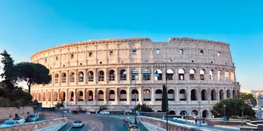 The Colosseum in Rome bathed in sunlight, with a clear blue sky, cars parked nearby, and a few people walking along the streets.