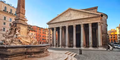 Historic stone building with large columns and a triangular pediment, adjacent to a stone fountain with sculpted figures.