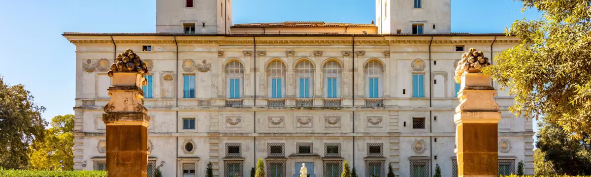 A grand historic villa with two tall towers, surrounded by manicured gardens, statues, and large columns under a clear blue sky.